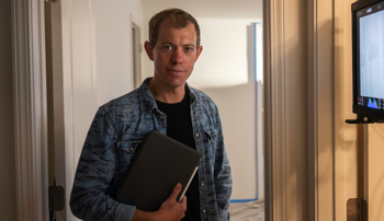 Brock Hanson wearing a denim shirt holds a notebook while standing in a softly lit hallway, looking at the camera.