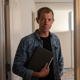 Brock Hanson wearing a denim shirt holds a notebook while standing in a softly lit hallway, looking at the camera.