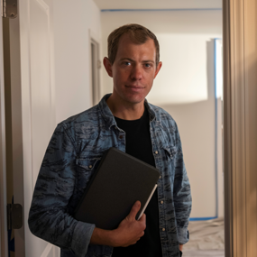 Brock Hanson wearing a denim shirt holds a notebook while standing in a softly lit hallway, looking at the camera.