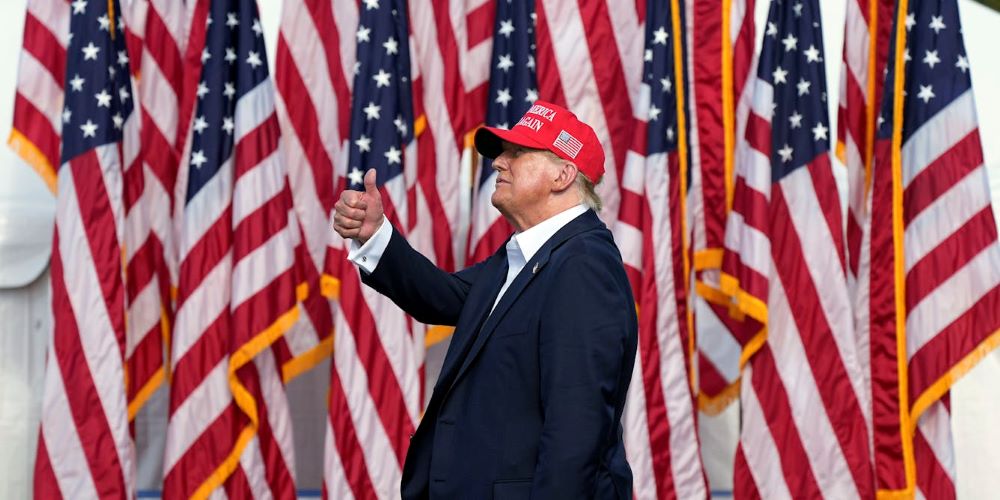 Trump giving a thumbs up in front of American flags and wearing a red MAGA hat