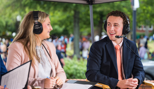 Two students wearing headsets talk to each other while broadcasting a parade