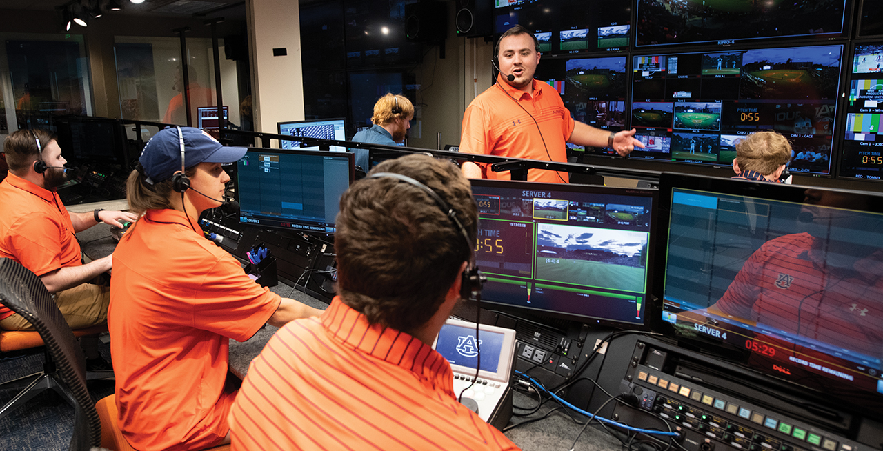 A group of people wearing orange shirts working in a broadcast control room with multiple screens displaying various camera feeds.