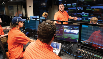 A group of people wearing orange shirts working in a broadcast control room with multiple screens displaying various camera feeds.