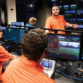 A group of people wearing orange shirts working in a broadcast control room with multiple screens displaying various camera feeds.