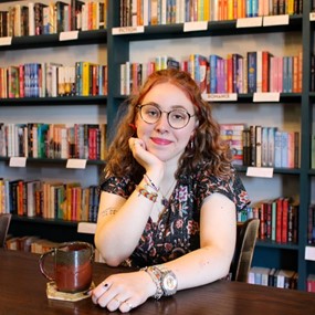 Louise Schulmann-Darsy sits at a cafe with a bookshelf of romance novels behind her
