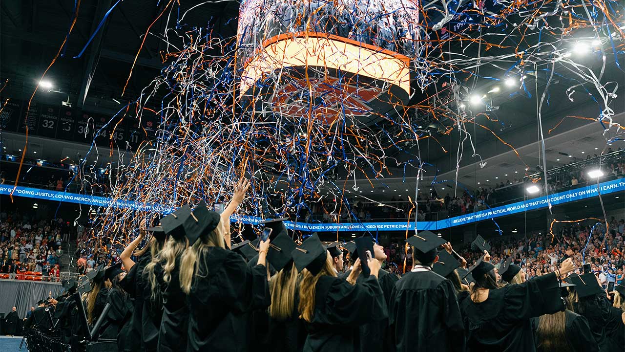 Graduates in caps and gowns reaching for blue and orange confetti at graduation