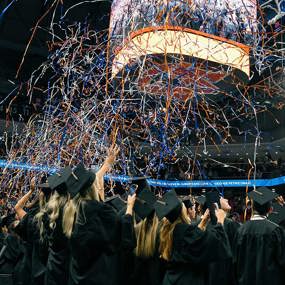 Graduates in caps and gowns reaching for blue and orange confetti at graduation