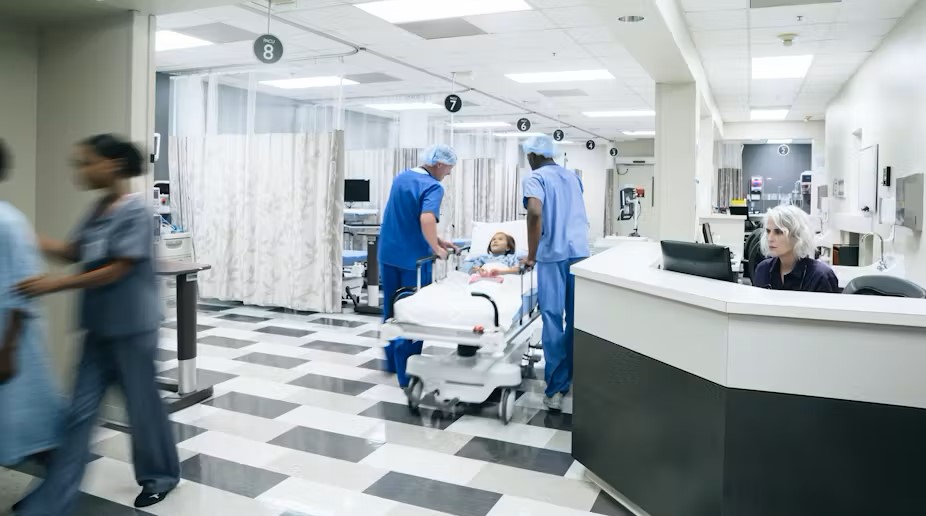 Near a nurses' station in a busy hospital, staff make the rounds and a patient in a gurney is wheeled down the hallway.