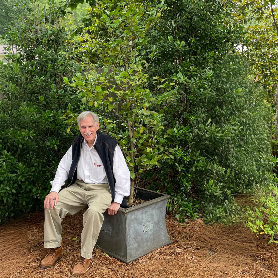 An older man with a mustache, wearing a white shirt, beige pants, a dark vest, and brown shoes, sits on the edge of a large square planter. The planter contains a young tree surrounded by lush greenery and pine straw in an outdoor garden setting.