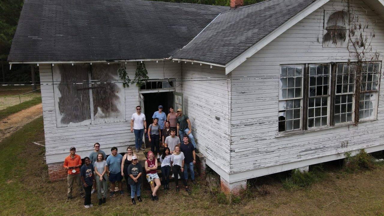Photo of a dilapidated school house with a crew of people posing for a photo