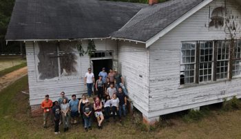 Photo of a dilapidated school house with a crew of people posing for a photo