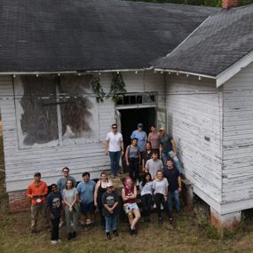Photo of a dilapidated school house with a crew of people posing for a photo