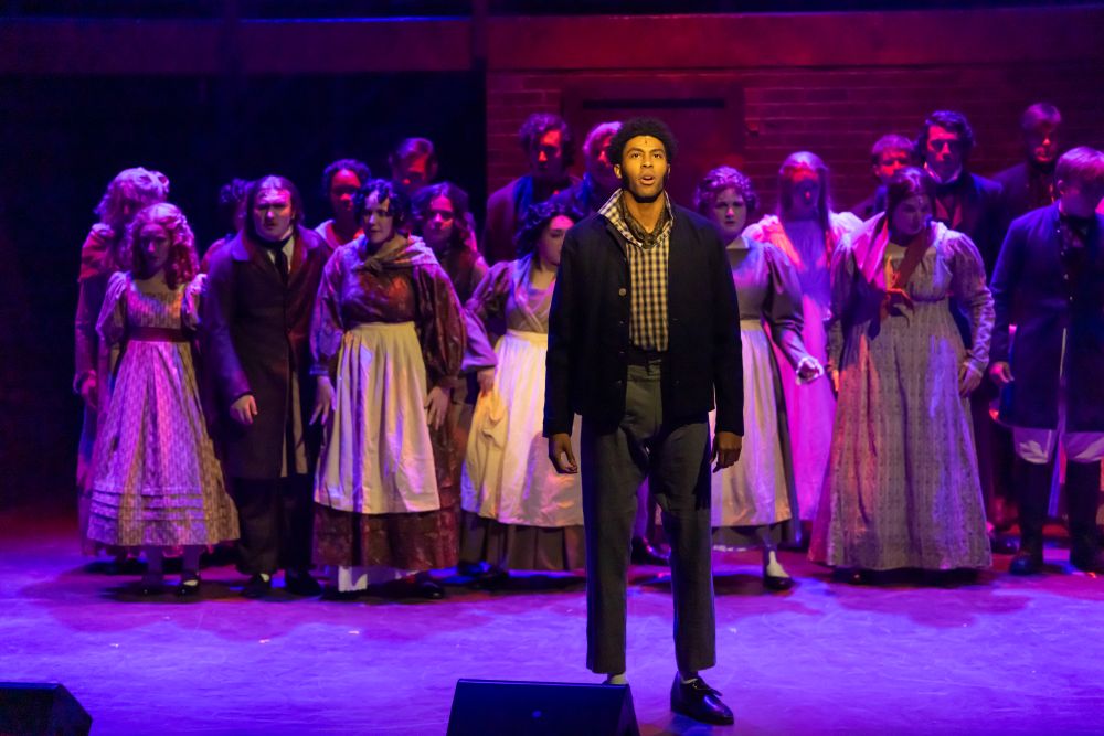 A student actor stands center stage with the “Sweeney Todd” ensemble gathered behind him under purple and red stage lighting.