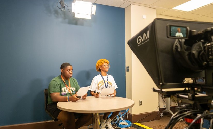 two students sitting at a table in front of a camera pretending to be reporters