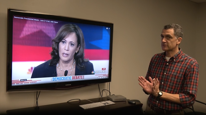 Mike Milford speaking in front of a television screen showing Harris debate footage