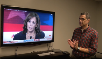 Mike Milford speaking in front of a television screen showing Harris debate footage