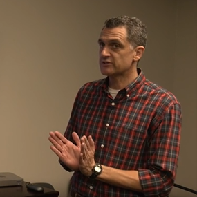 Mike Milford speaking in front of a television screen showing Harris debate footage