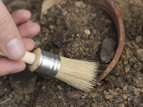 Hand holding a brush sweeping dirt away from a piece of pottery in the ground