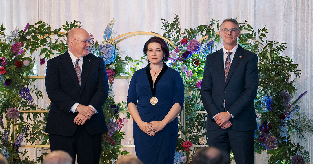 Emily stands proudly wearing a medal while President Roberts and Dean Hicks smile for the photo