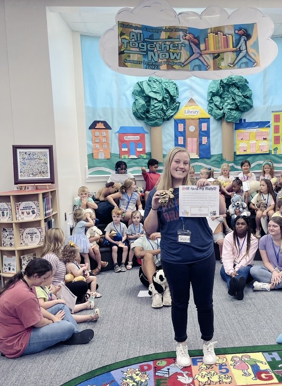 a student stands in front of children assembled for story time at the local library