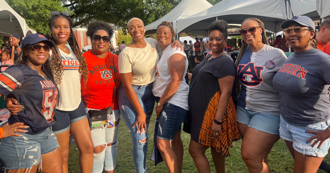 A group of women wearing Auburn shirts and casual clothing pose together outdoors during an Auburn campus event, with white tents and trees in the background.