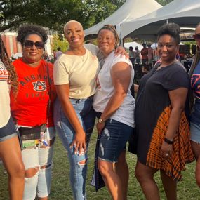 A group of women wearing Auburn shirts and casual clothing pose together outdoors during an Auburn campus event, with white tents and trees in the background.
