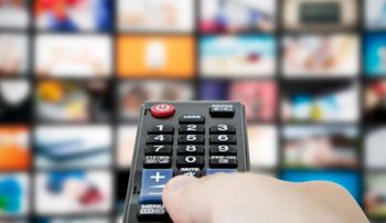 A hand pressing buttons on a remote in front of a screen showing various video streams