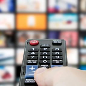 A hand pressing buttons on a remote in front of a screen showing various video streams