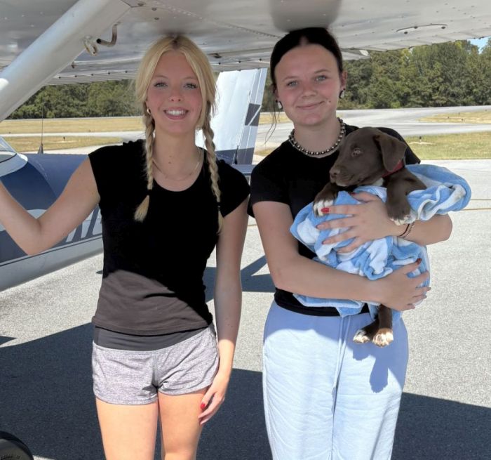 Two women standing under a plane's wing, one is holding a puppy wrapped in small blanket.