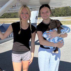 Two women standing under a plane's wing, one is holding a puppy wrapped in small blanket.