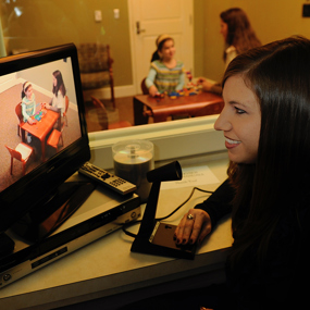A therapist watches an adult and child build blocks on a TV screen while the same child and adult can be seen through a one-way mirror in the background.