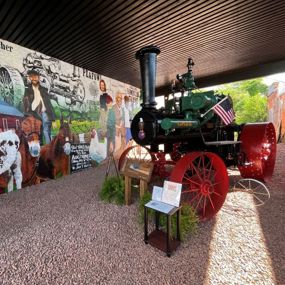 Steam tractor standing in pavilion where agricultural collage mural is displayed on wall