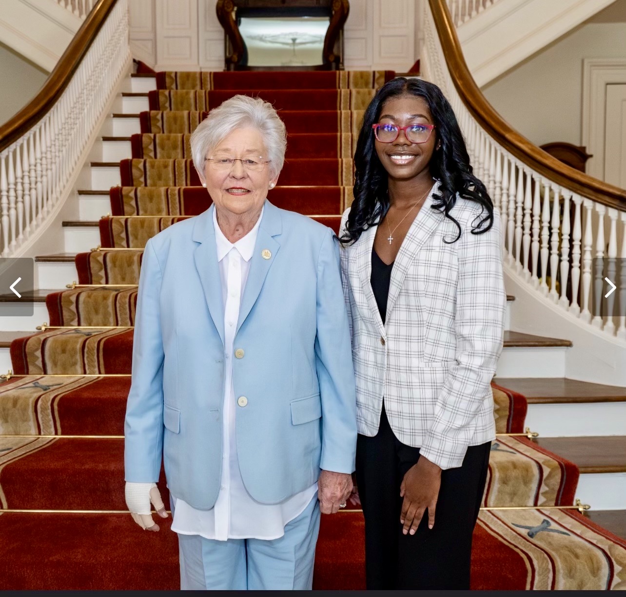 Kasia Nicholson stands proudly next to Governor Kay Ivey in front of a red velvet staircase