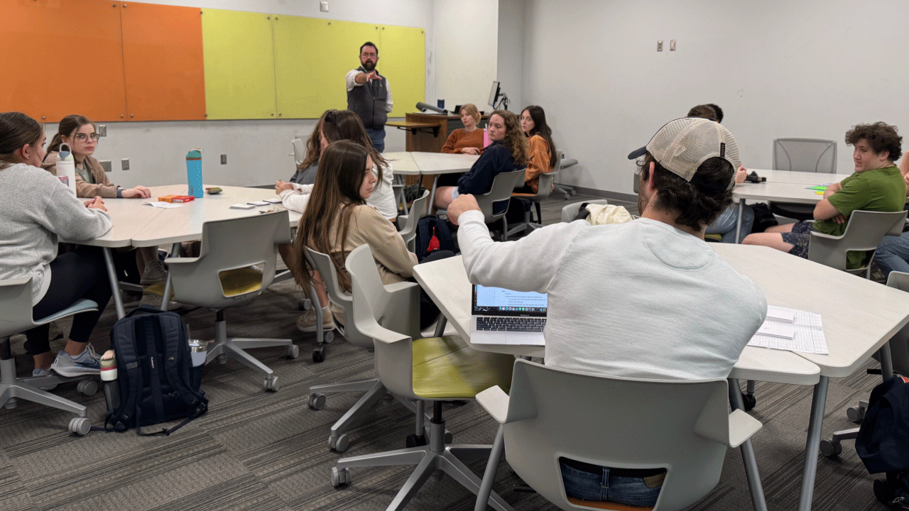 A classroom of college students sits in small groups at circular tables while an instructor at the front of the room gestures toward the class. Several students are engaged in discussion, some taking notes or working on laptops. Colorful boards hang on the front wall, and backpacks and water bottles are scattered around the room.