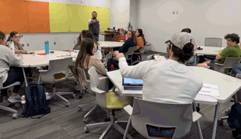 A classroom of college students sits in small groups at circular tables while an instructor at the front of the room gestures toward the class. Several students are engaged in discussion, some taking notes or working on laptops. Colorful boards hang on the front wall, and backpacks and water bottles are scattered around the room.