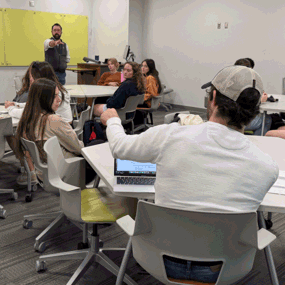 A classroom of college students sits in small groups at circular tables while an instructor at the front of the room gestures toward the class. Several students are engaged in discussion, some taking notes or working on laptops. Colorful boards hang on the front wall, and backpacks and water bottles are scattered around the room.
