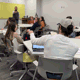 A classroom of college students sits in small groups at circular tables while an instructor at the front of the room gestures toward the class. Several students are engaged in discussion, some taking notes or working on laptops. Colorful boards hang on the front wall, and backpacks and water bottles are scattered around the room.