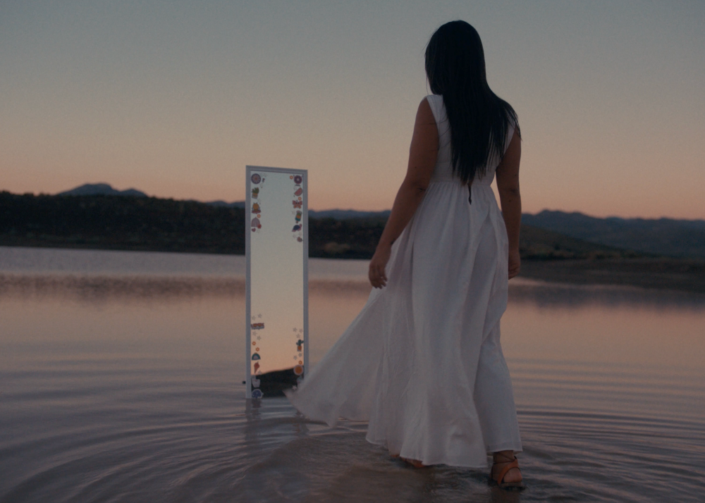 A woman in a long white dress standing in water at sunrise staring at a children&#x27;s mirror