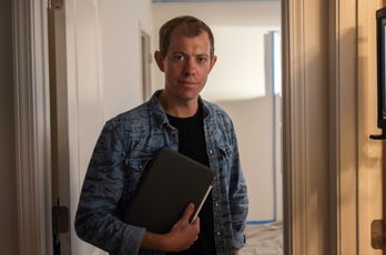 Brock Hanson wearing a denim shirt holds a notebook while standing in a softly lit hallway, looking at the camera.