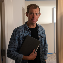 Brock Hanson wearing a denim shirt holds a notebook while standing in a softly lit hallway, looking at the camera.
