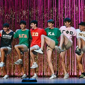 A row of young men in sorority t-shirts performing a kick line on stage