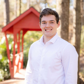 Biomedical sciences sophomore John Walker stands outdoors on Auburn University’s campus near a small red footbridge, wearing a white button-down shirt and smiling toward the camera