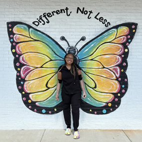 Celeste, who wears prosthetic arms, stands in front of a brick wall painted with butterfly wings