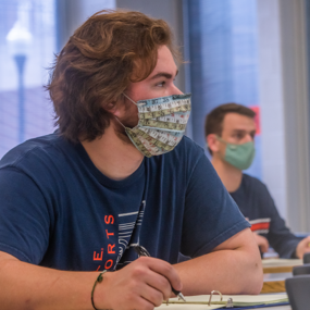 Two students wear face masks in a classroom with text reading COVID plus speech clarity, the masked speaker