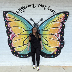 Celeste, who wears prosthetic arms, stands in front of a brick wall painted with butterfly wings