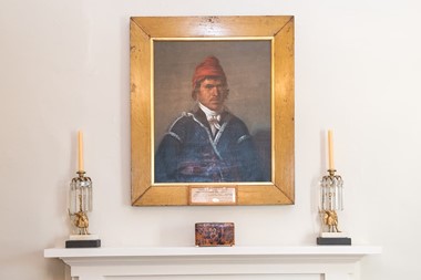 A mantle with a portrait of a Native American above it. He wears a red turban and blue jacket. Candlesticks sit on either side of the portrait. 